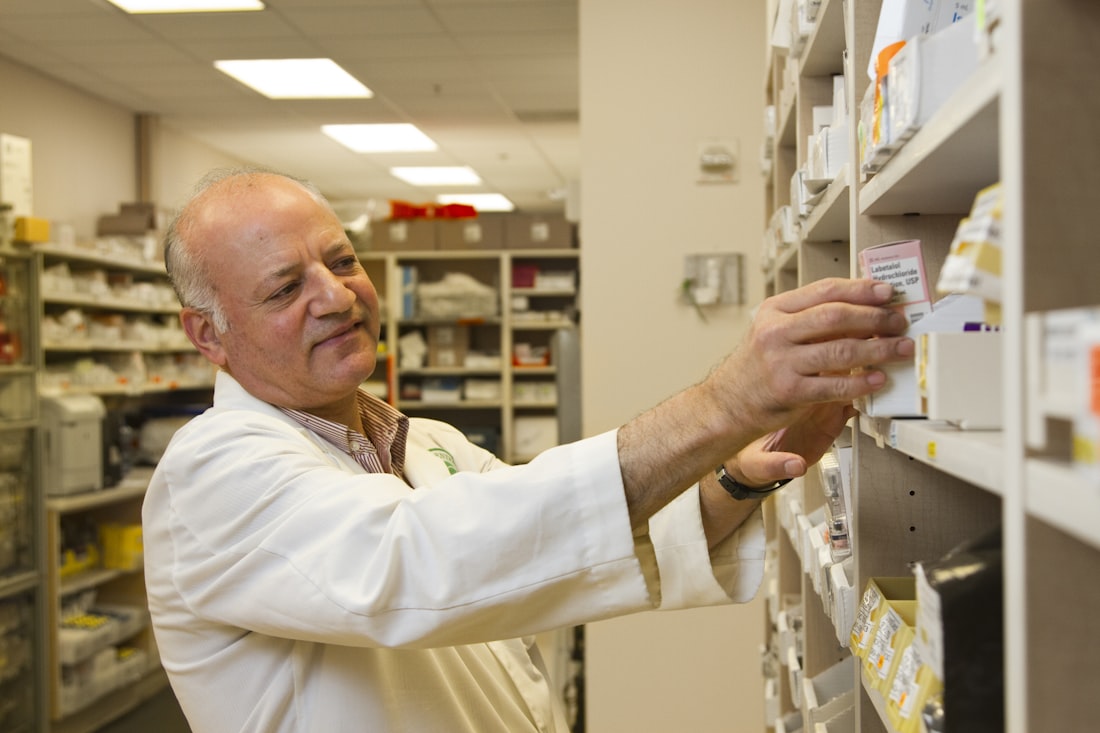 A pharmacist in a white coat selecting a medication package from labelled shelving.