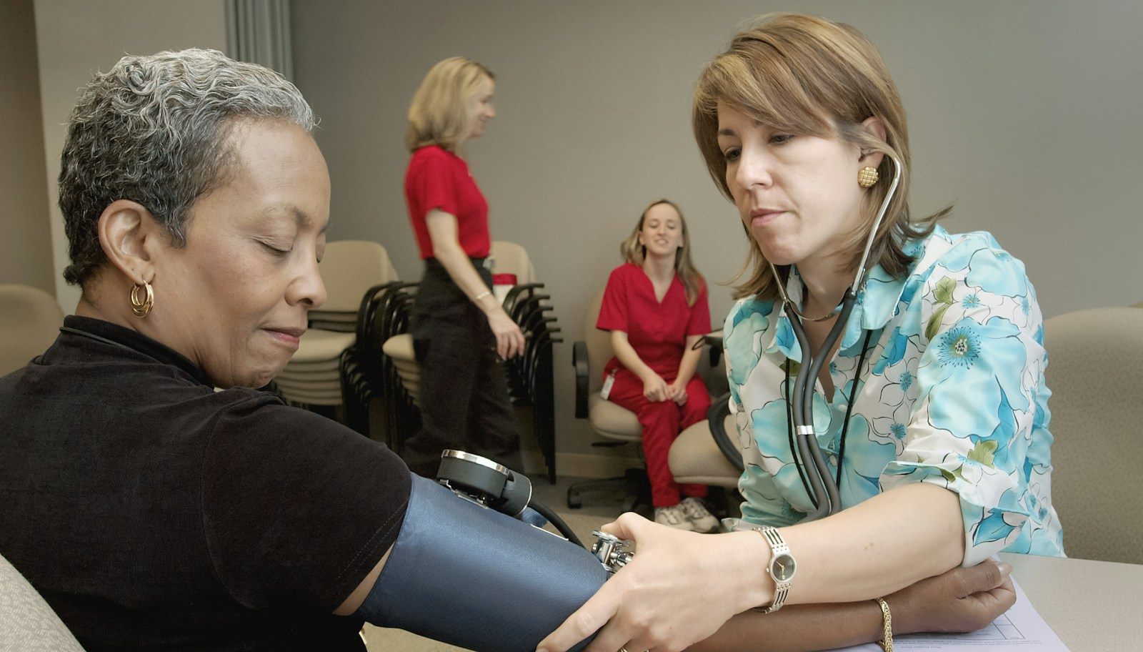 A nurse taking a seated patient's blood pressure during a health screening appointment, with other staff and waiting patients in the background.